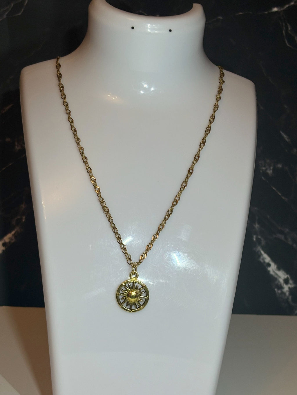 A gold-toned chain necklace with a circular pendant displayed on a white mannequin bust against a dark marbled background.
