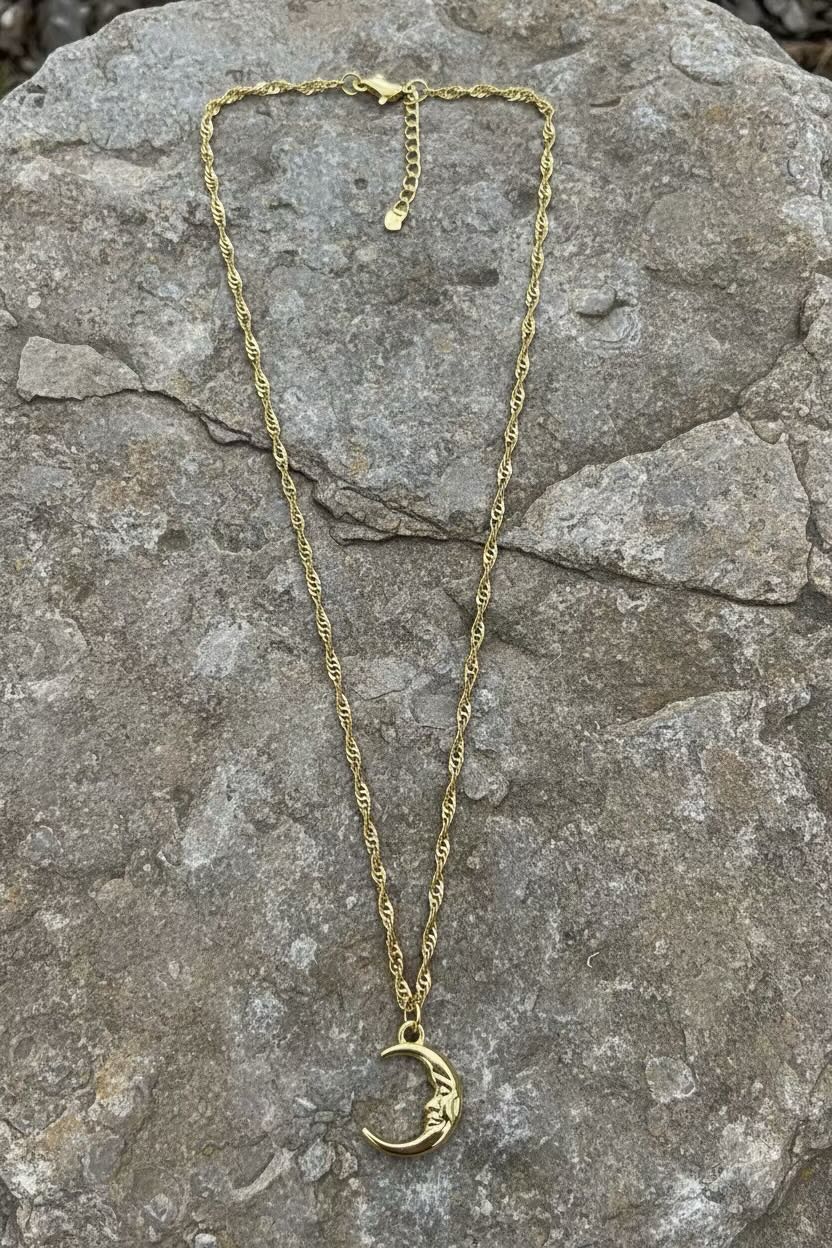 A gold necklace with a crescent moon face pendant featuring a small face, displayed on a textured gray rock background.