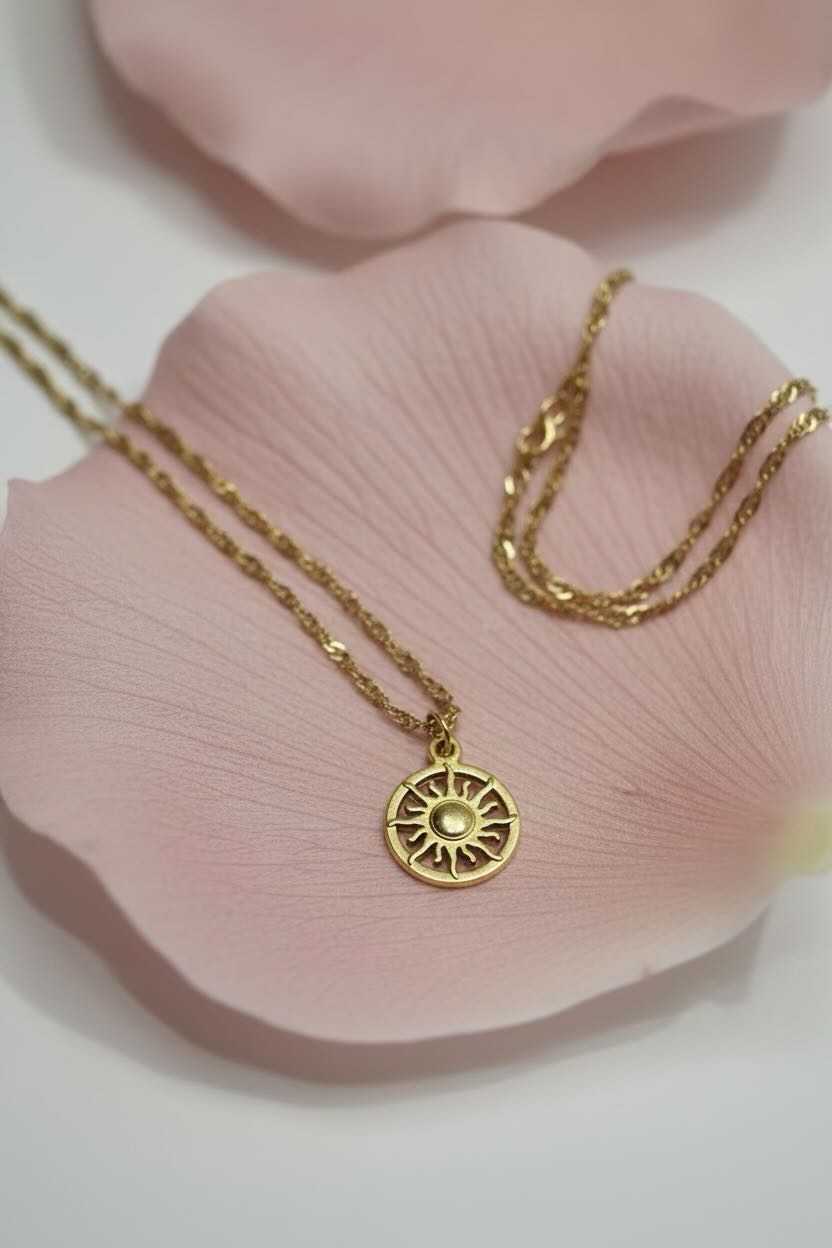 A gold necklace with a sunburst pendant displayed on a soft pink rose petal against a white background.