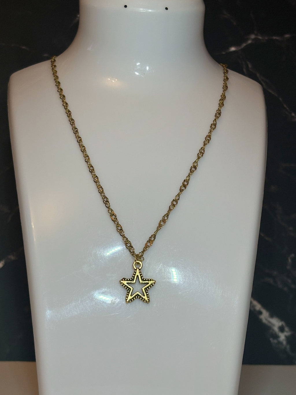 A gold-tone star pendant necklace displayed on a glossy white mannequin bust against a dark marble-patterned background.