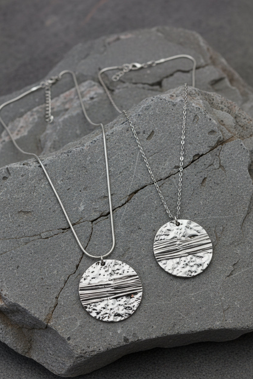 Two silver pendant necklaces with textured circular pendants resting on a gray stone slab.