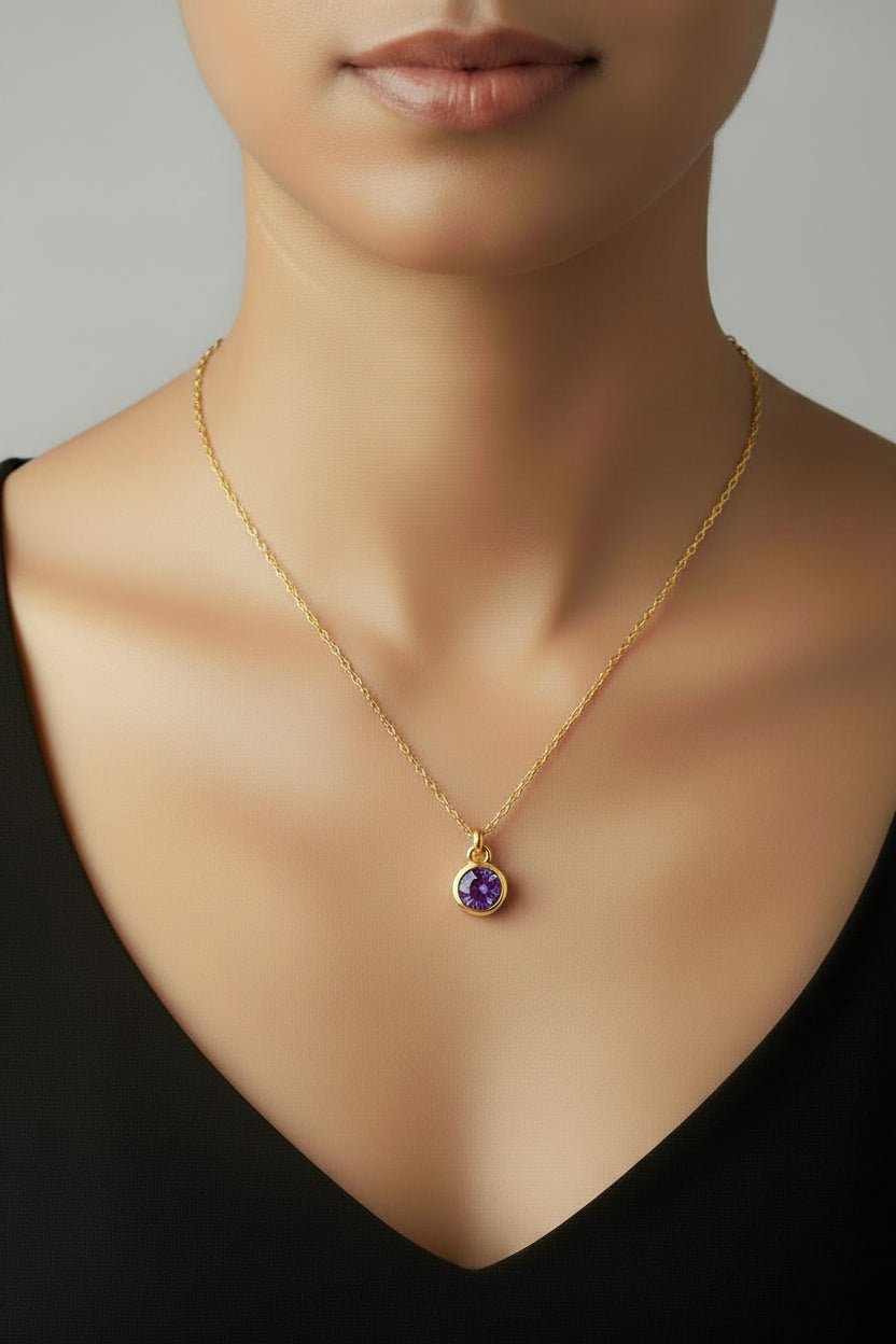 A close-up of a model's neck and upper chest wearing a delicate gold chain necklace with a small oval pendant featuring a purple gemstone surrounded by a gold halo.