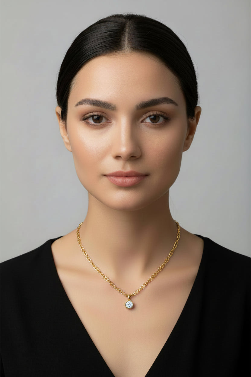 A woman with dark hair pulled back wears a delicate gold chain necklace with a small round clear gemstone pendant, photographed against a plain light gray background.