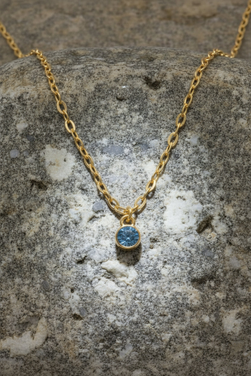 A gold chain necklace with a small round blue gemstone pendant, displayed on a textured gray stone surface.