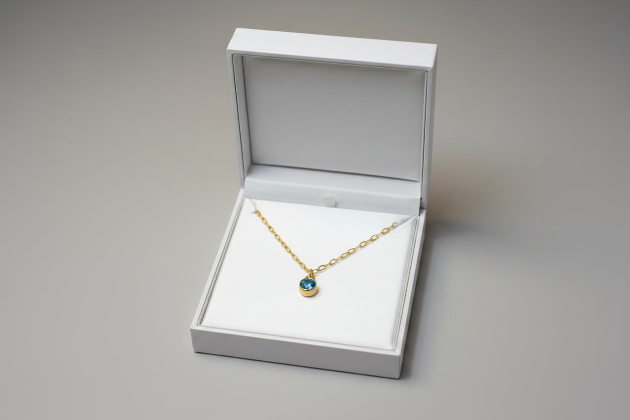 A gold chain necklace with a small oval blue gemstone pendant, displayed inside an open white jewelry box on a light gray background.