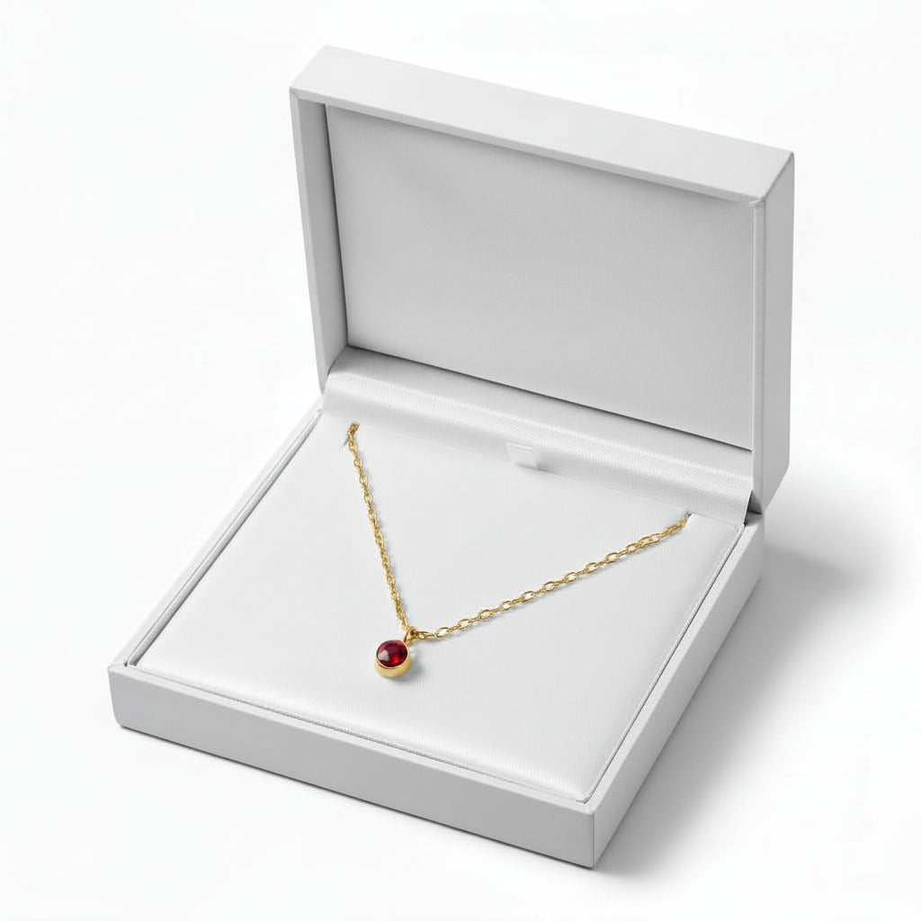 A gold chain necklace with a small red oval pendant displayed inside an open white jewelry box on a clean white background.