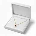 A gold chain necklace with a small red oval pendant displayed inside an open white jewelry box on a clean white background.