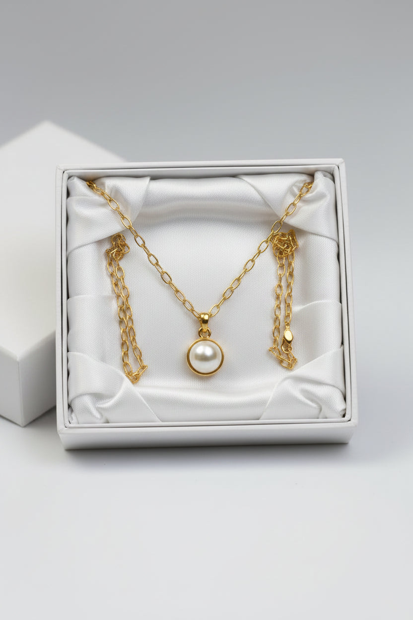 A gold chain necklace with a single round white pearl pendant, presented in an open white gift box lined with white satin, on a light gray background.