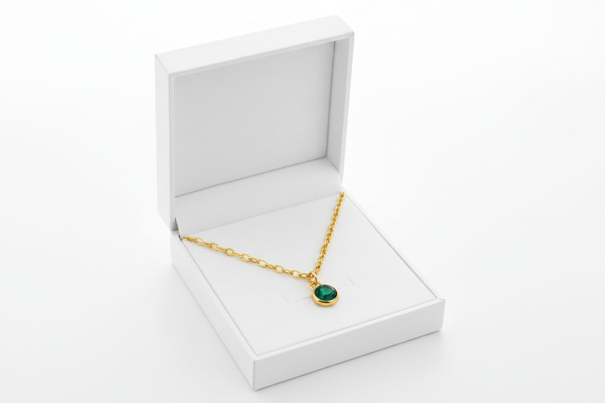 A gold chain necklace with a small oval green gemstone pendant, presented inside an open white jewelry box on a clean white background.