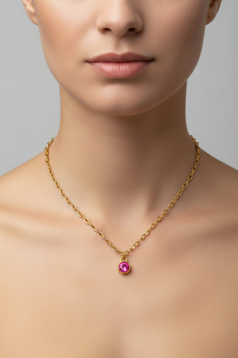 A close-up of a model's neck and upper chest wearing a gold-toned chain necklace with a small teardrop-shaped pink gemstone pendant.