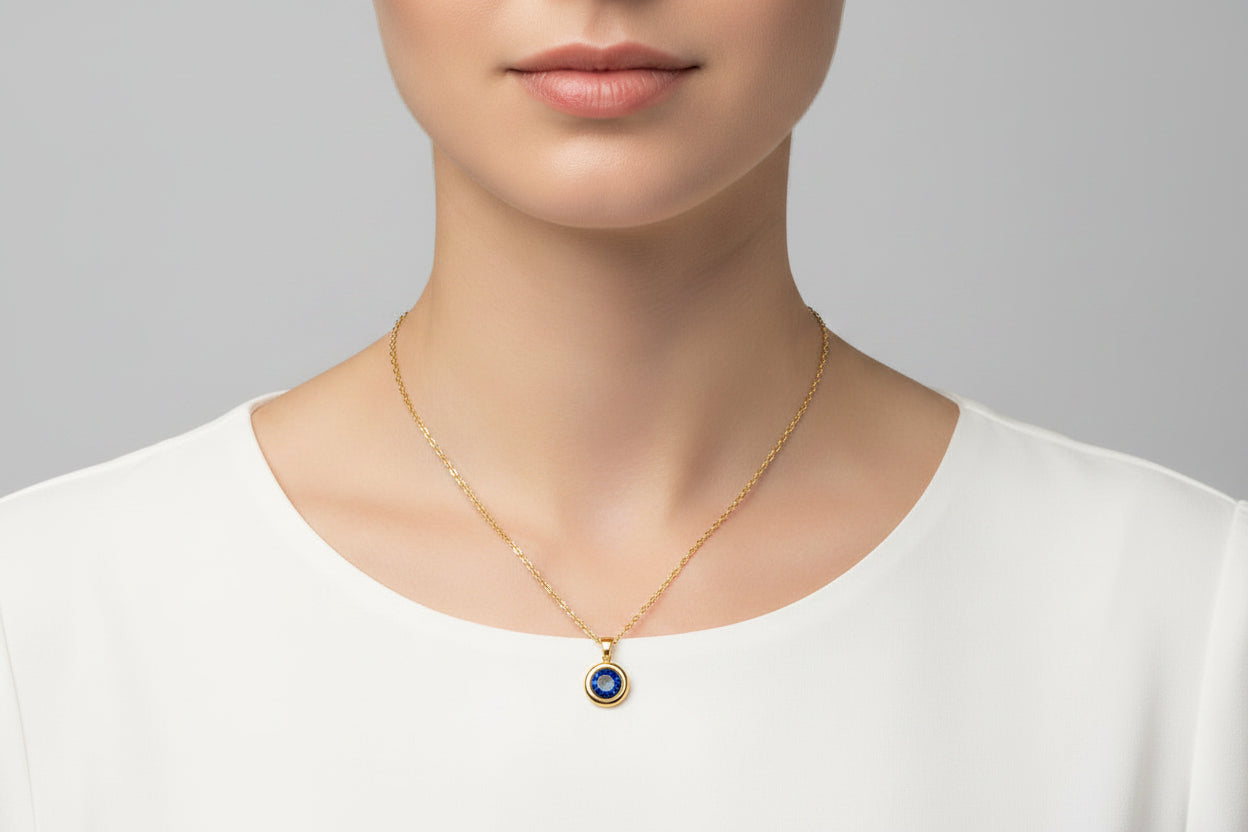 A close-up of a model wearing a delicate gold chain necklace with a small evil eye pendant featuring blue and white details, set against a neutral gray background.