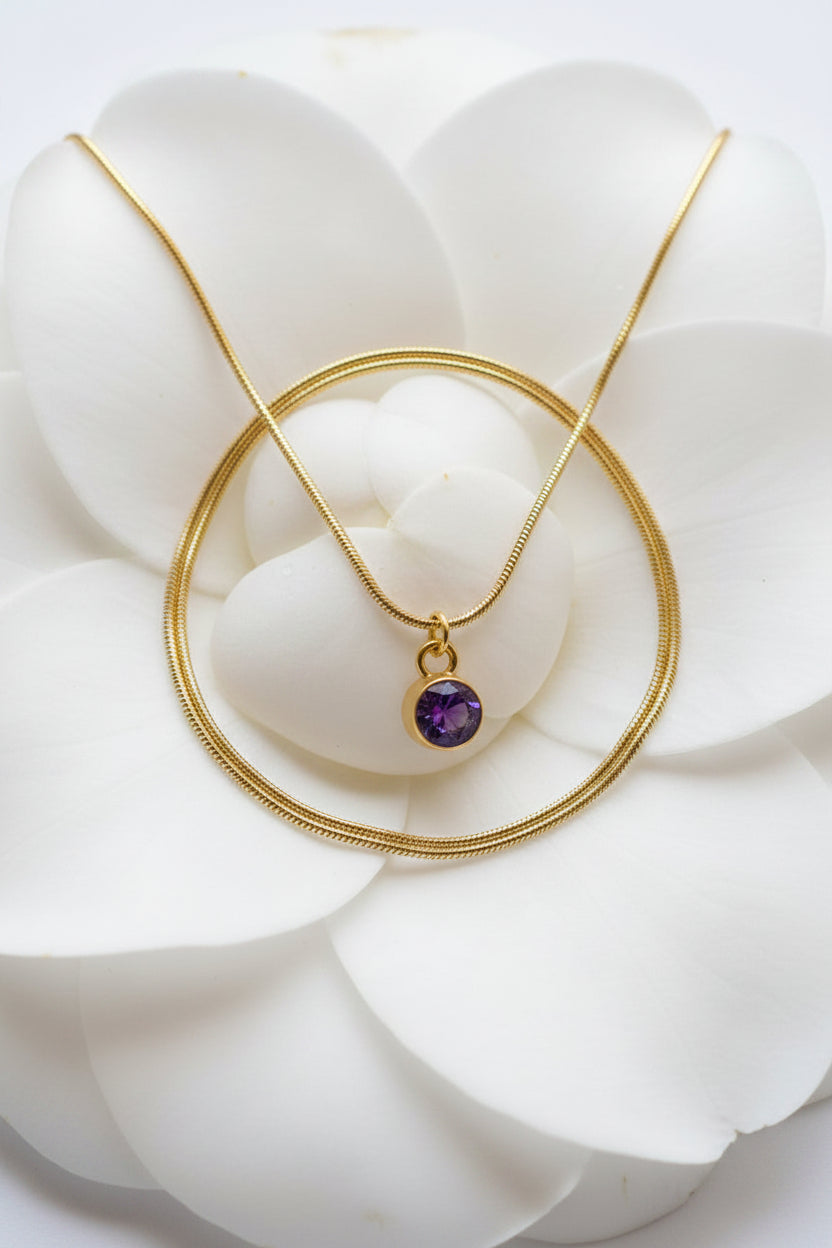 A gold chain necklace with a small oval purple gemstone pendant, displayed on a large white flower.