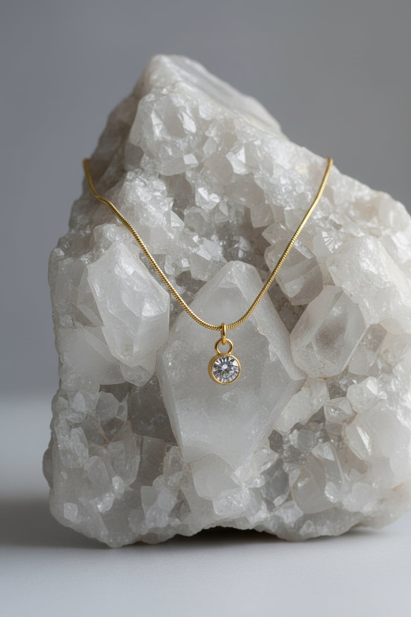 A delicate gold chain necklace with a round bezel-set clear gemstone pendant, displayed draped over a large white crystal geode specimen against a neutral gray background.