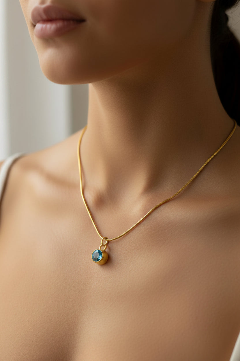 A close-up of a model wearing a delicate gold chain necklace with a small oval pendant featuring a light blue stone.