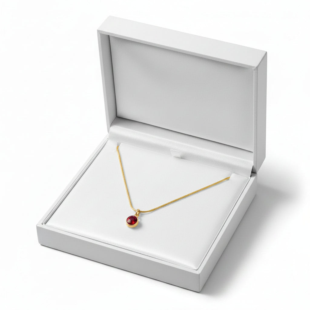 A gold necklace with a small red oval pendant displayed inside an open white jewelry box on a clean white background.