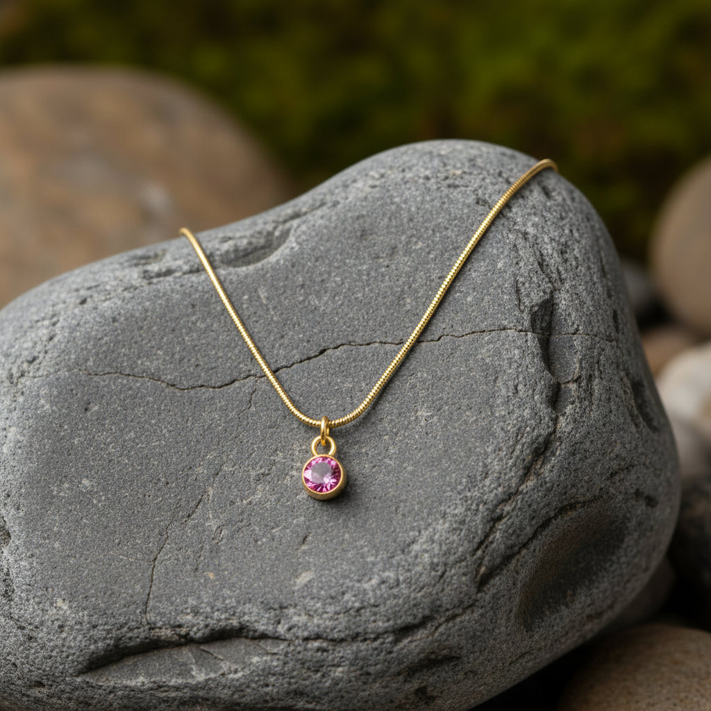 A gold snake chain necklace with a small teardrop-shaped pink gemstone pendant, displayed on a textured gray river stone with a blurred natural background.