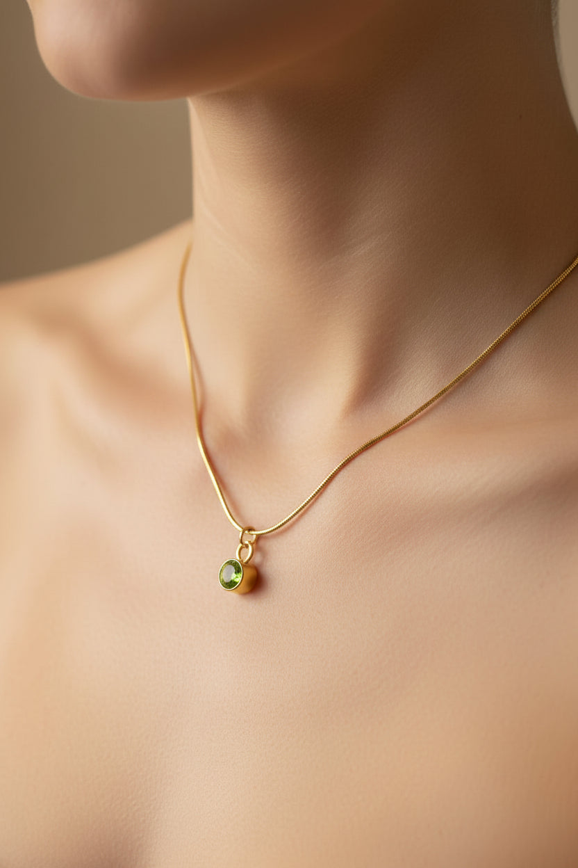 A delicate gold chain necklace with a small green oval pendant, shown worn on a model's neck against a soft beige background.