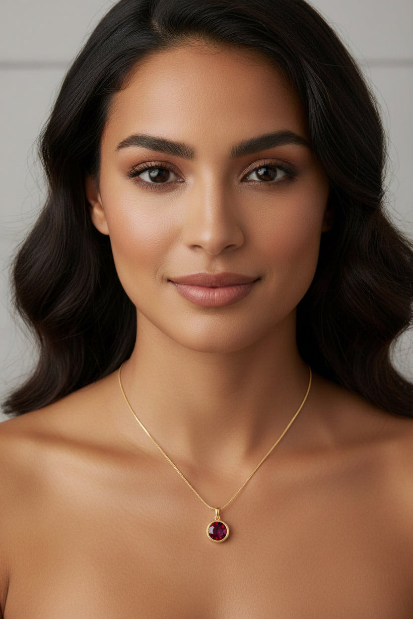 A close-up portrait of a woman wearing a delicate gold chain necklace with a round dark red gemstone pendant, set against a soft light gray background.