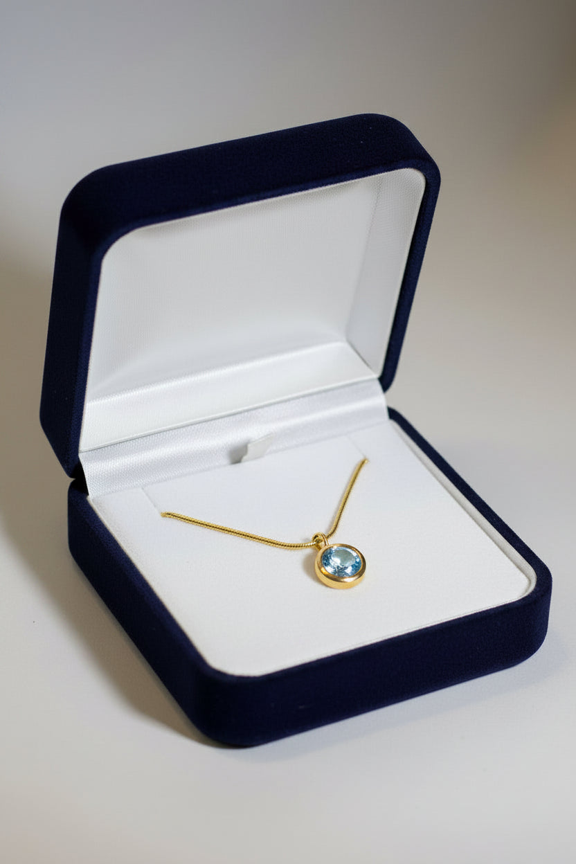 A gold necklace with a small oval blue gemstone pendant, displayed inside an open dark blue velvet jewelry box with a white interior.