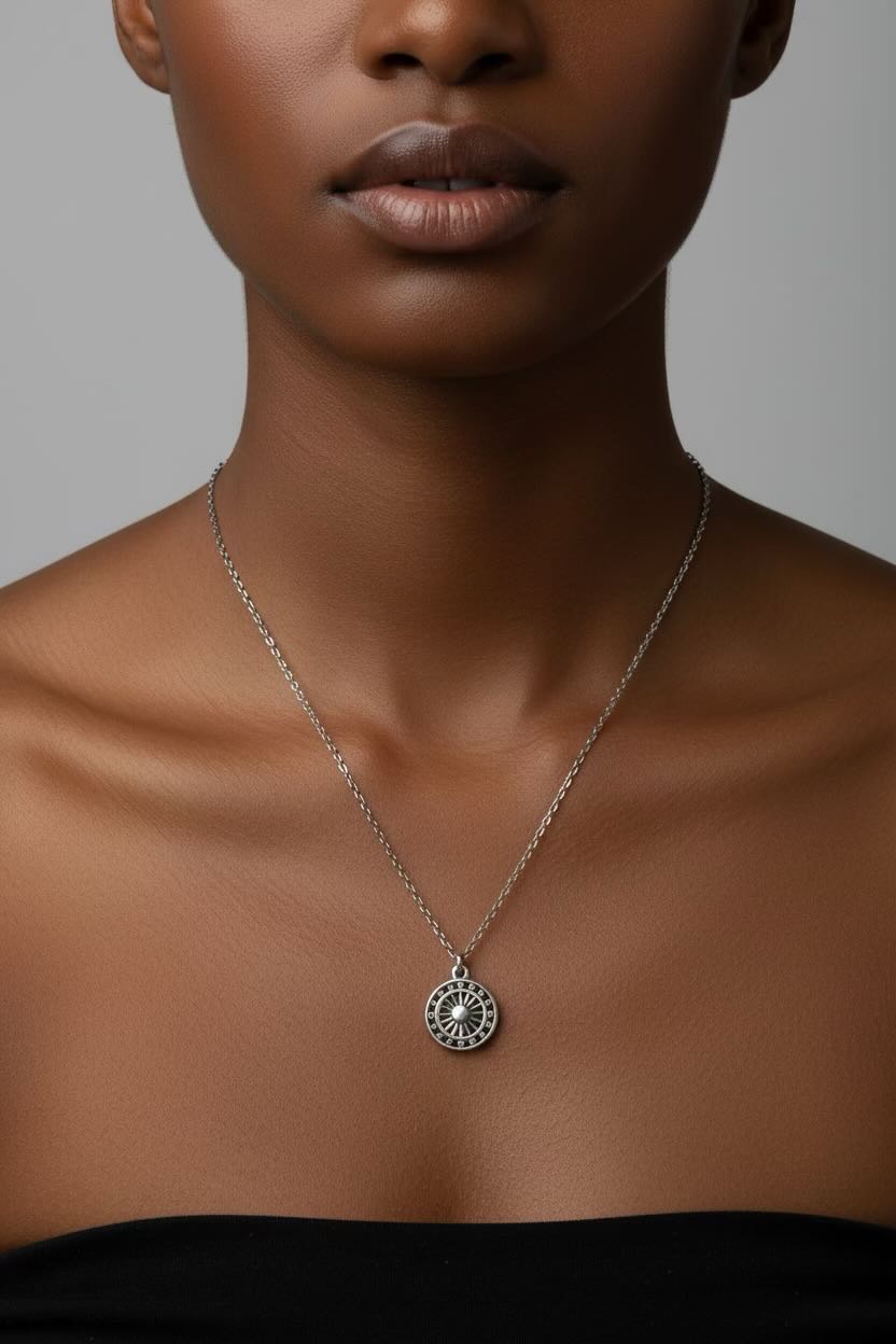 A close-up of a model wearing a delicate silver chain necklace with a circular element coin pendant, set against a neutral gray background.