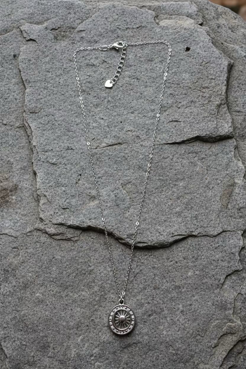 A silver necklace with a circular element coin pendant and a small dangling tag, displayed on a textured gray stone surface.