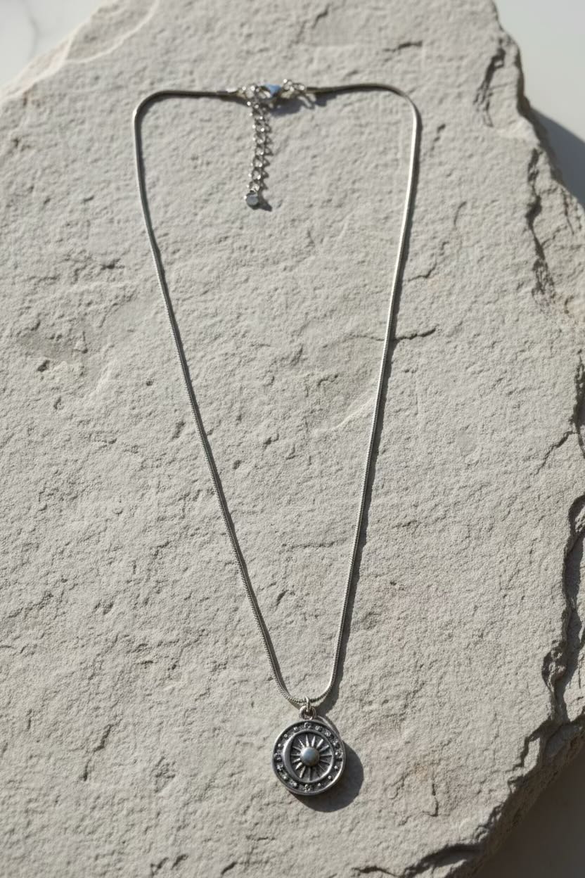 A silver-toned necklace with a circular element coin pendant, displayed on a textured light gray stone surface.