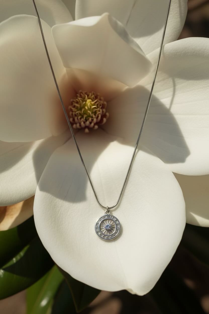 A silver-toned round pendant necklace with a element design hangs from a delicate chain, presented on the center of a large white magnolia flower.