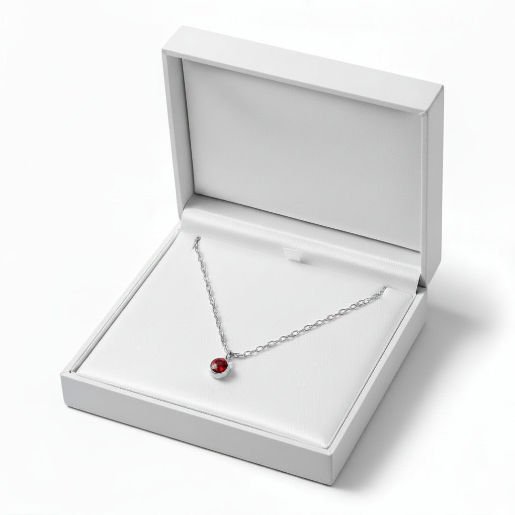 A silver necklace with a small red birthstone pendant displayed inside an open white jewelry box on a clean white background.
