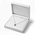 A silver necklace with a small red birthstone pendant displayed inside an open white jewelry box on a clean white background.