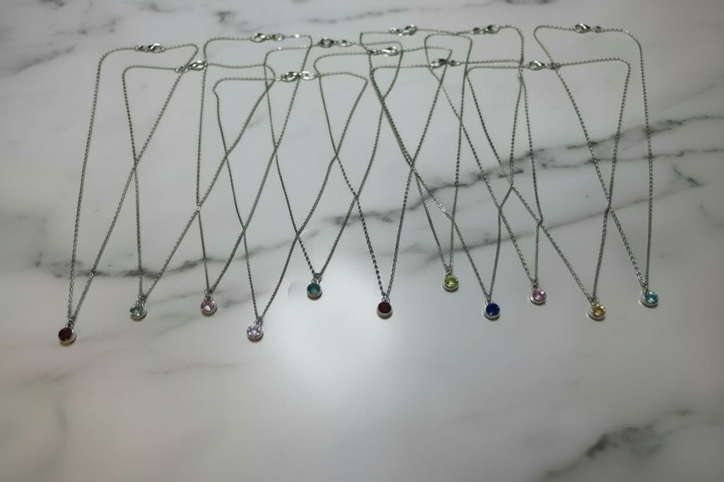 A set of eleven silver chain necklaces with round birthstone pendants in various colors, arranged on a plain white surface.