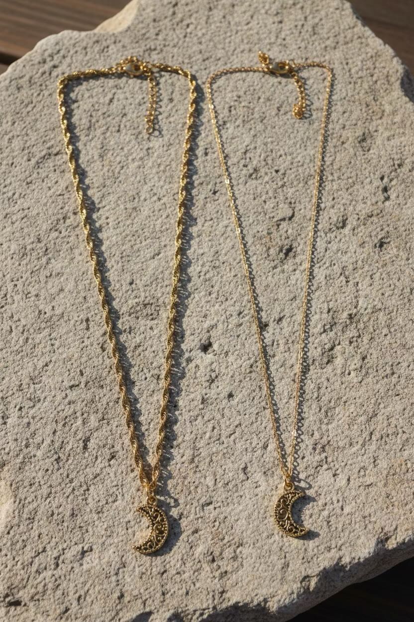 Two gold crescent moon necklaces with chain extenders, displayed on a textured light gray stone surface.