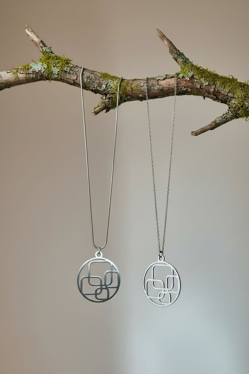 Two silver geometric pendant necklaces hanging from a mossy tree branch against a soft beige background.