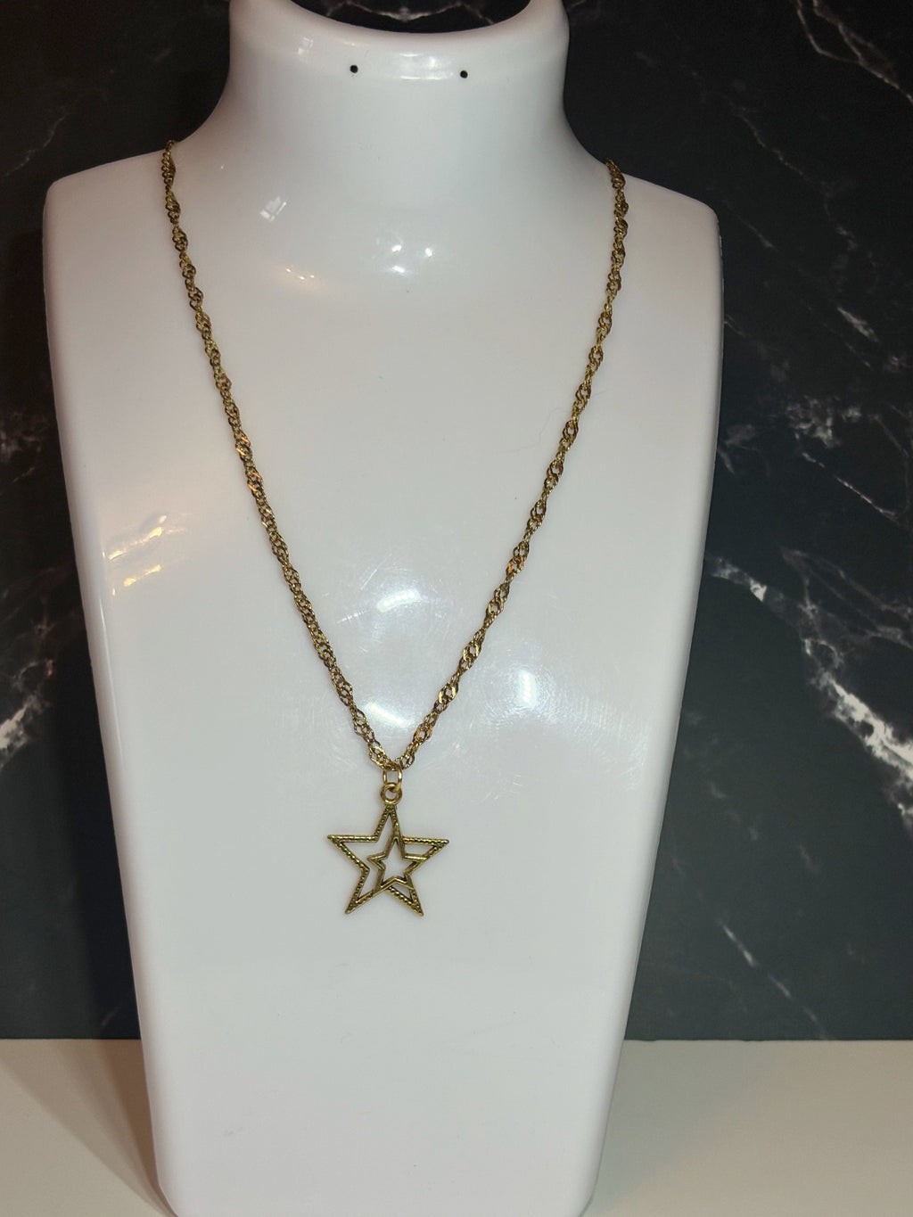A gold-toned chain necklace with an open star-shaped pendant, displayed on a white jewelry bust against a dark marble-patterned background.