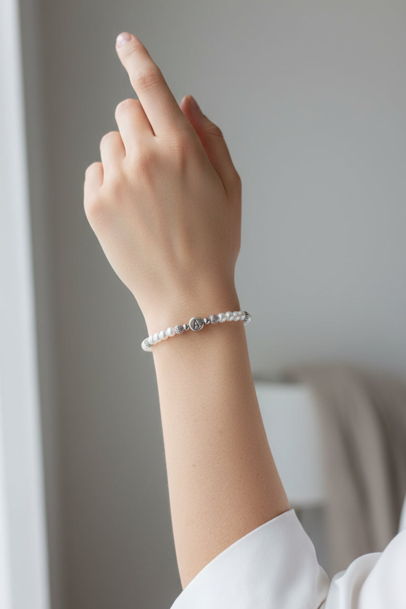 A beaded stretch bracelet displayed on a white mannequin arm. The bracelet features white glass pearls, small silver spacer beads, and a central round silver bead engraved with the letter A.