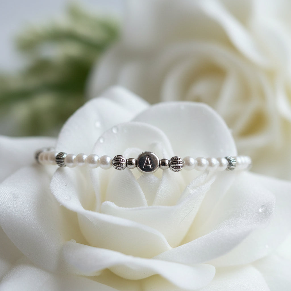 A delicate beaded bracelet with white glass pearls and silver spacer beads, featuring a central silver disc engraved with the letter 'A', displayed on a white petal.