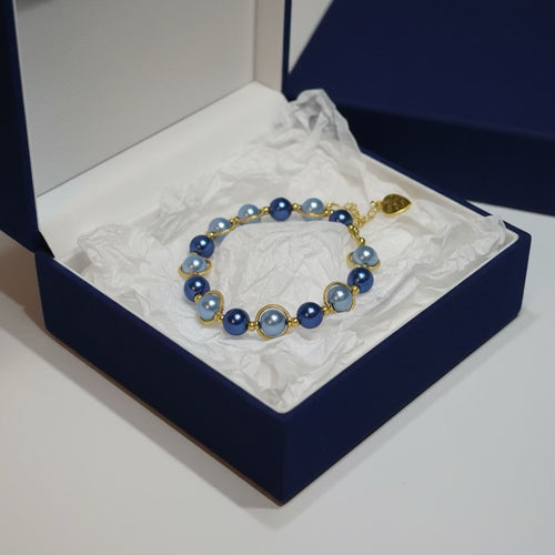 A pearl bracelet with dark royal blue and dark baby blue glass pearls and gold-toned settings, displayed inside an open navy blue jewelry box with white tissue paper.