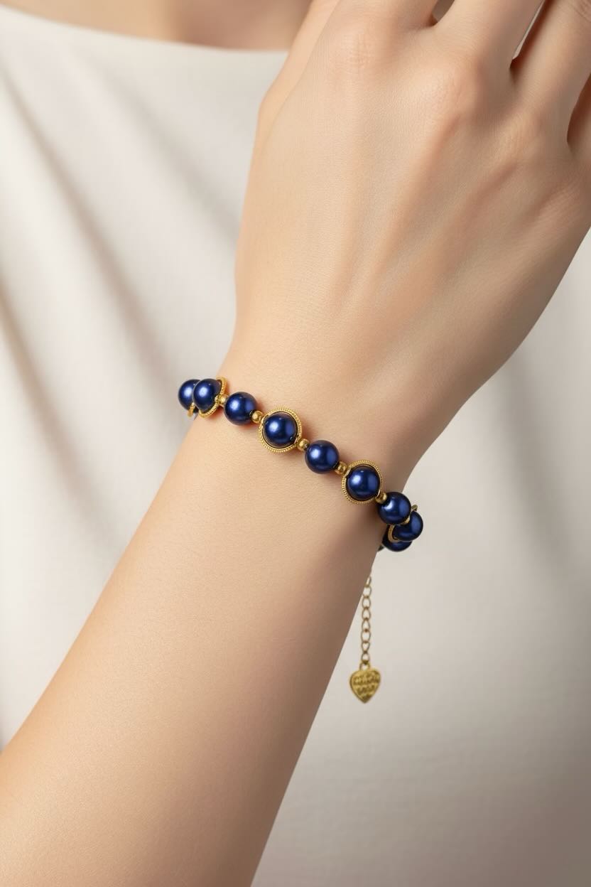 A close-up of a person's wrist wearing a delicate bracelet made of dark royal blue pearls and gold-toned metal links, with a small dangling gold heart charm.