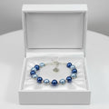 A beaded bracelet with blue and dark baby blue pearls and silver-toned metal links, displayed inside an open white jewelry box on a white surface.