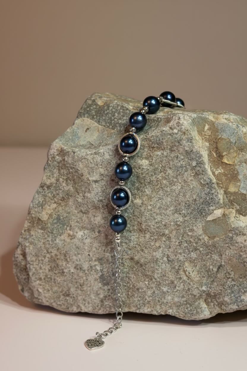 A beaded bracelet with steel blue pearls and silver spacer beads draped over a textured gray stone, featuring a silver chain extender with a small heart charm.