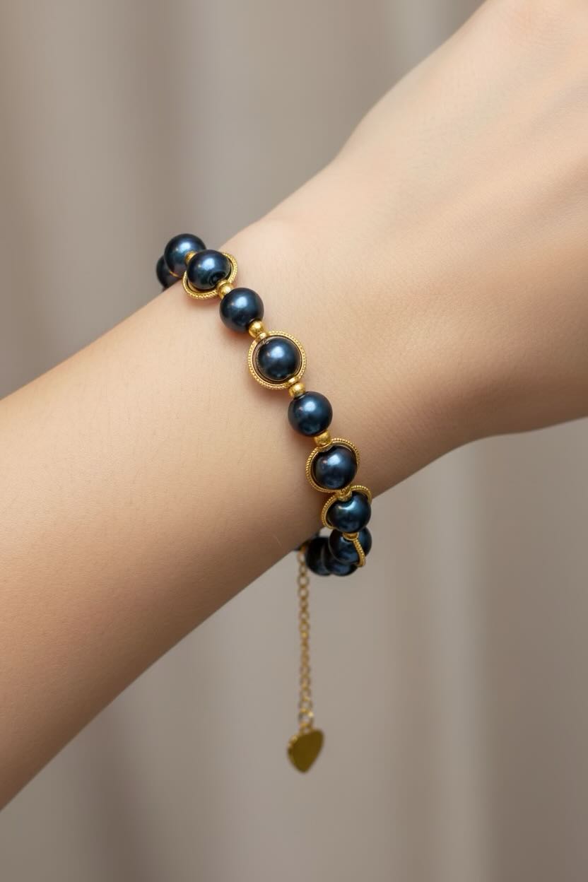 A close-up of a person's wrist wearing a bracelet made of  steel blue glass pearls and gold-toned settings, with a small dangling gold heart charm.