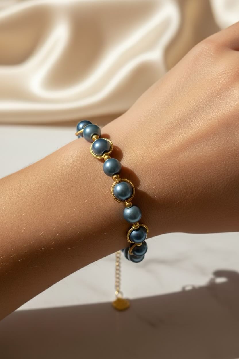 A close-up of a person's wrist wearing a delicate bracelet made of round teal blue glass pearls and gold spacer rings, with a small gold chain extender and charm hanging below. The background features draped satin fabric.